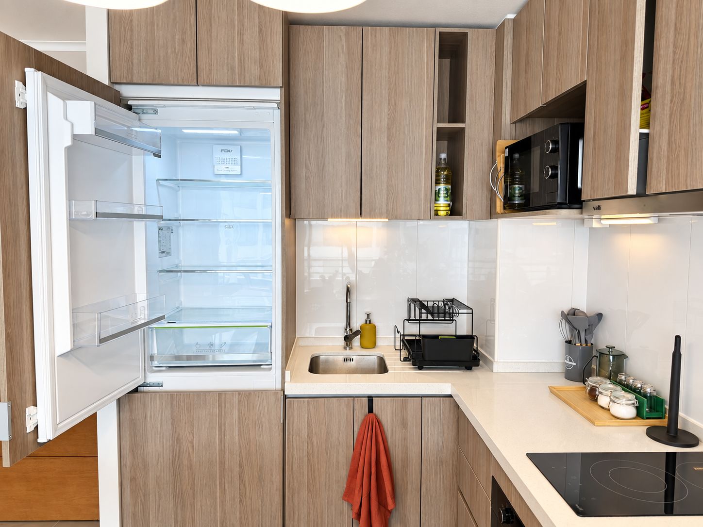 Una cocina moderna con armarios de madera, horno empotrado, pileta de acero inoxidable y cocina por inducción negra.
