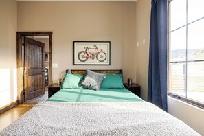 Ein Schlafzimmer mit einem Bett, einem gerahmten Kunstwerk mit einem Fahrradmotiv, einer Holztür und Blick durch ein Fenster.