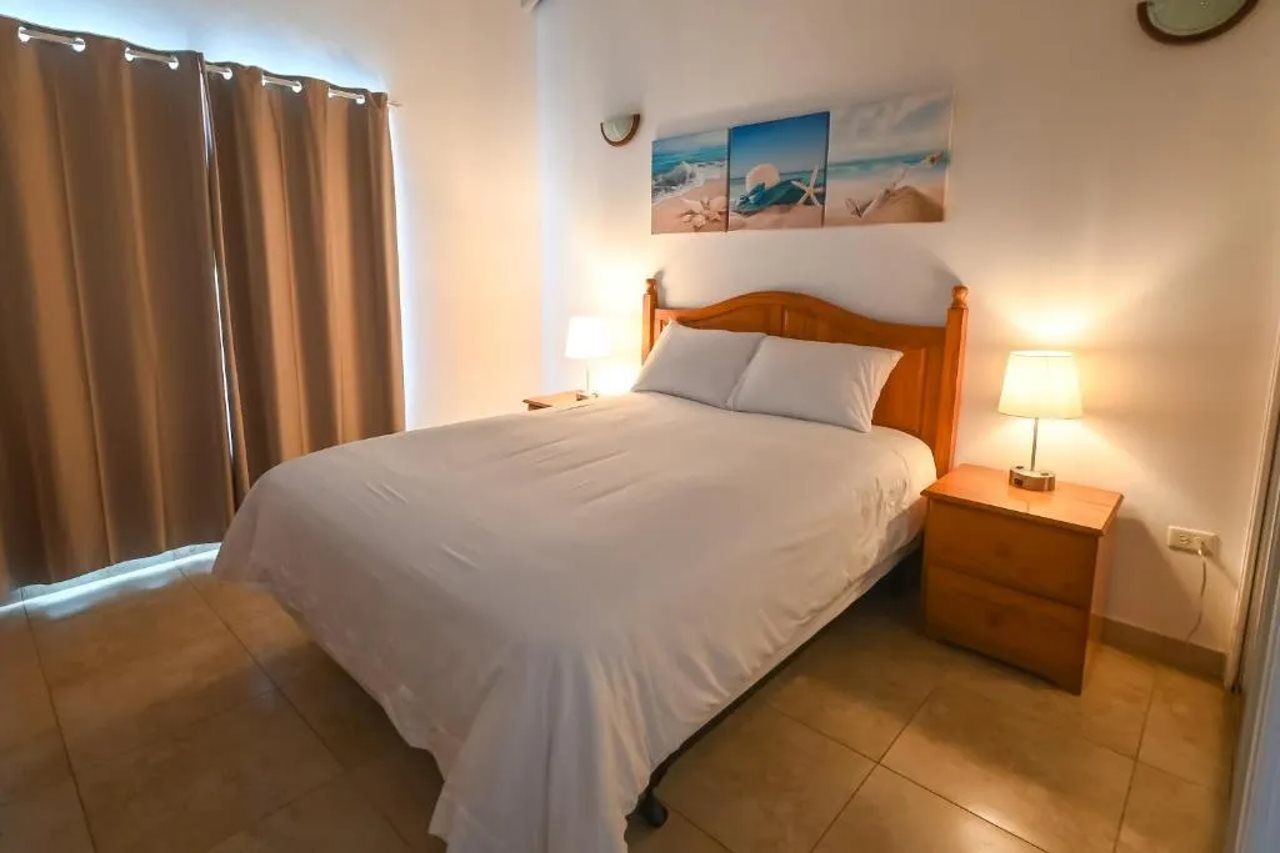 A bedroom with a bed, two bedside tables with lamps, a wall clock, and a window with curtains.