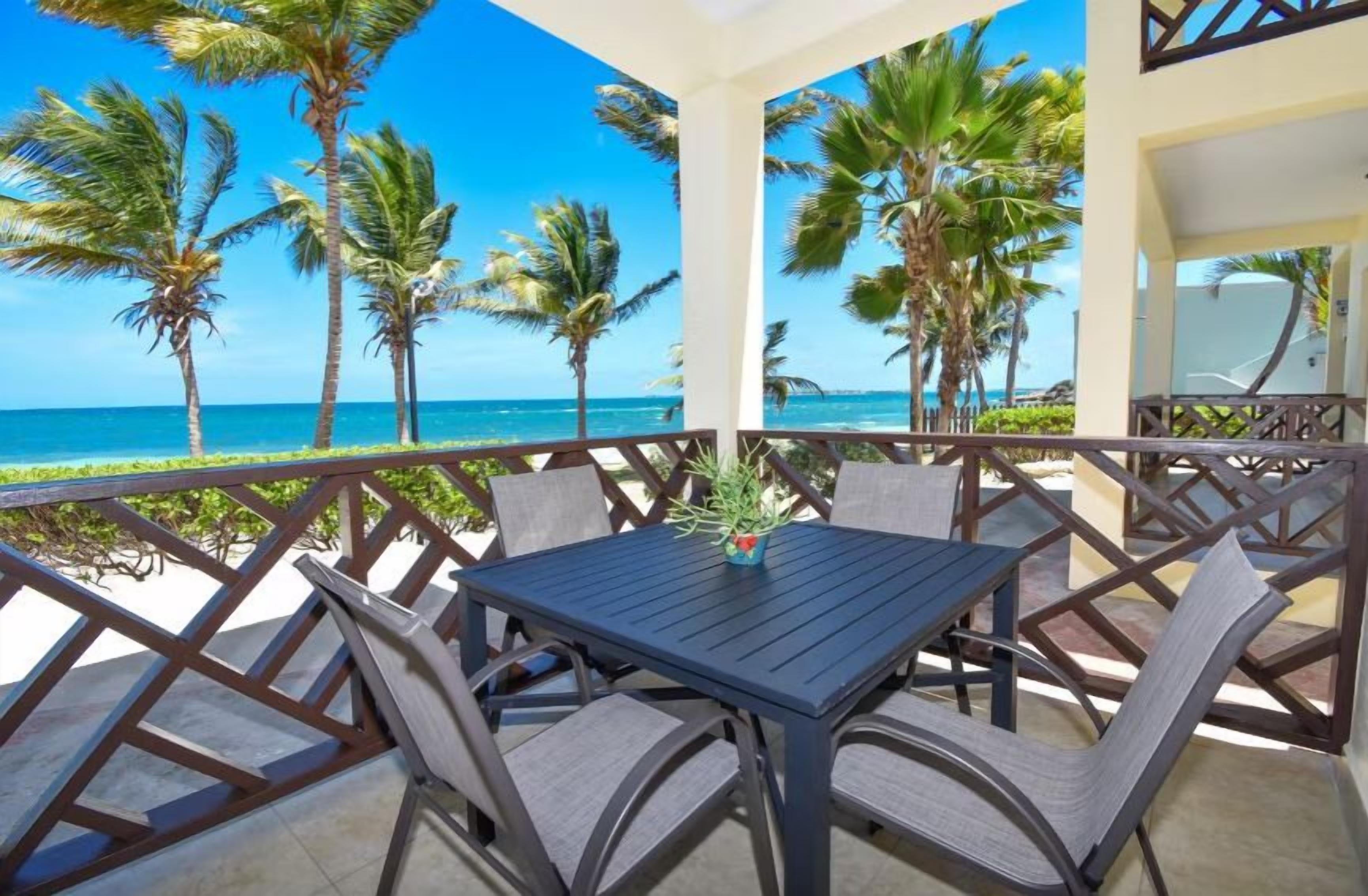 A balcony with a table and chairs, overlooking a beach with palm trees and the ocean.