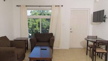 A room with a sofa, a small table, a dining table with chairs, a TV mounted on the wall, and a door.