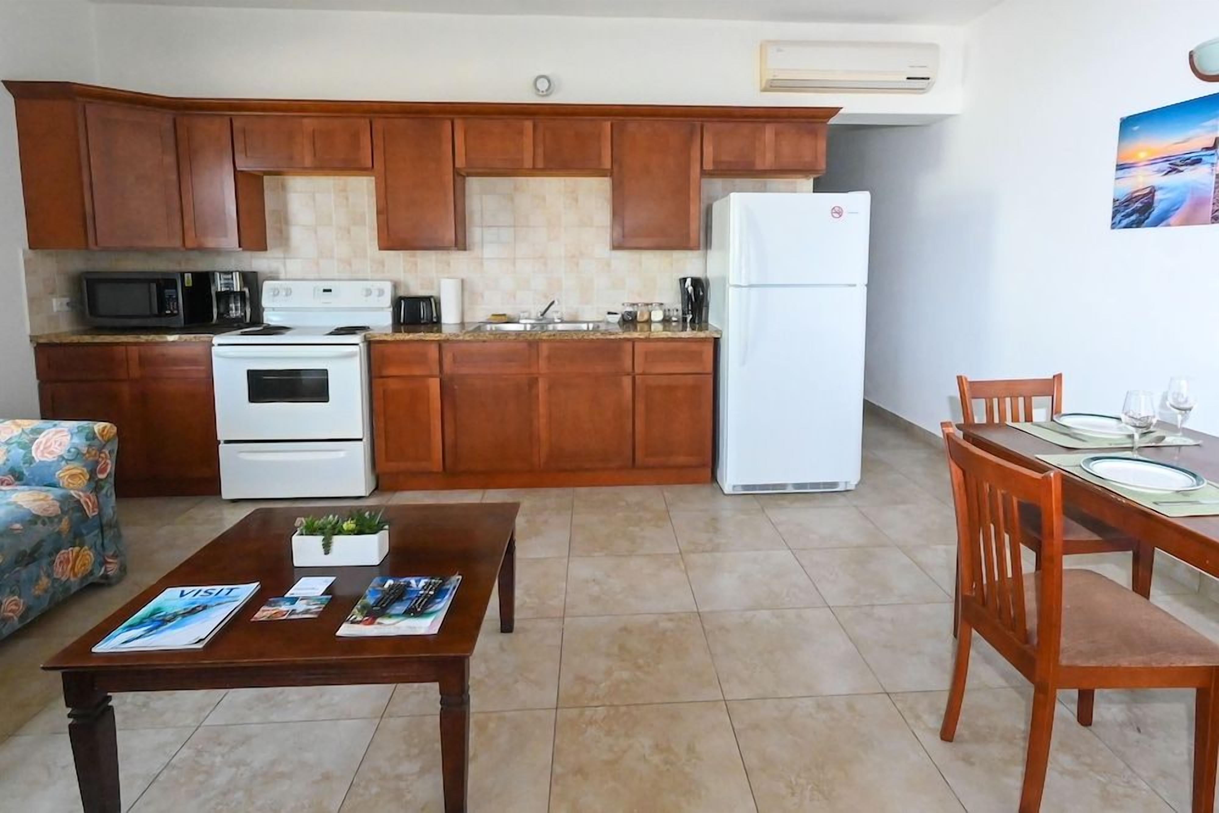 Una cocina compacta con armarios de madera, un refrigerador blanco, un microondas y una estufa. Hay una zona de comedor con una mesa y sillas, y una pequeña planta sobre la mesa.