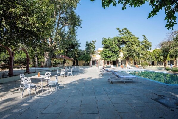 An outdoor patio area with tables and chairs, a pool, and a building in the background. - Justa Alwarbagh Sariska (Alwar)