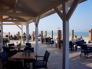 An outdoor dining area with wooden tables and chairs, overlooking a beach. - Holiday Home in Renesse With Swimming Pool (Renesse)