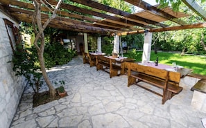 Een terras buiten met stenen vloer, houten tafels en banken, en een pergola.