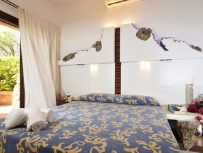 A bedroom with a bed, curtains, a bedside table with a lamp, and a wall with a unique rock-like design. - L"ea di Lavru - Apt 10 (Loiri Porto San Paolo)