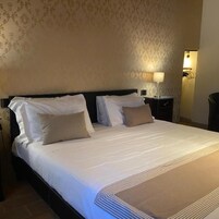 Una cama bien tendida con sábanas blancas y dos almohadas, una mesita de noche con una lámpara, y una pared con papel tapiz estampado.