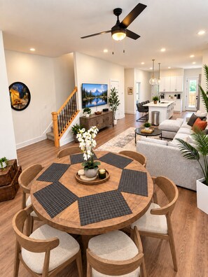 Een moderne woonkamer met een ronde houten tafel, comfortabele stoelen, een plafondventilator en een keuken op de achtergrond.