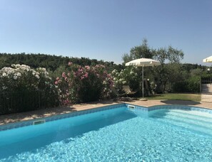 A swimming pool with a tiled edge, surrounded by a hedge with white and pink flowers, and a lawn area with a white umbrella. - Golf Maisonette Toscolano Maderno (Toscolano Maderno)