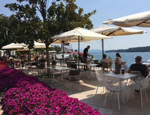 An outdoor café with tables and chairs under umbrellas, overlooking a body of water. - Golf Maisonette Toscolano Maderno (Toscolano Maderno)