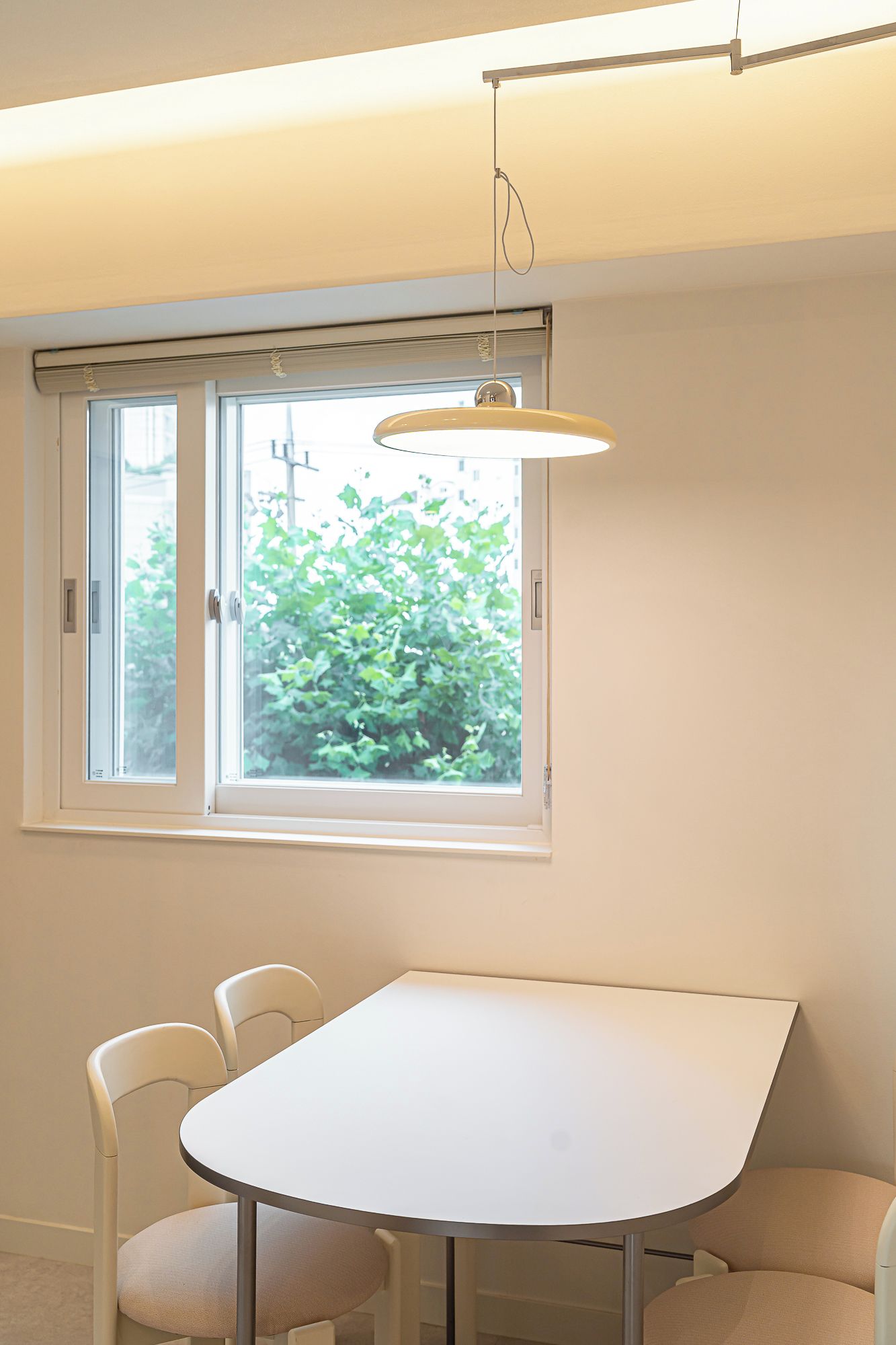 A room with a white table and two chairs, a window with a view of greenery, and a hanging light fixture.