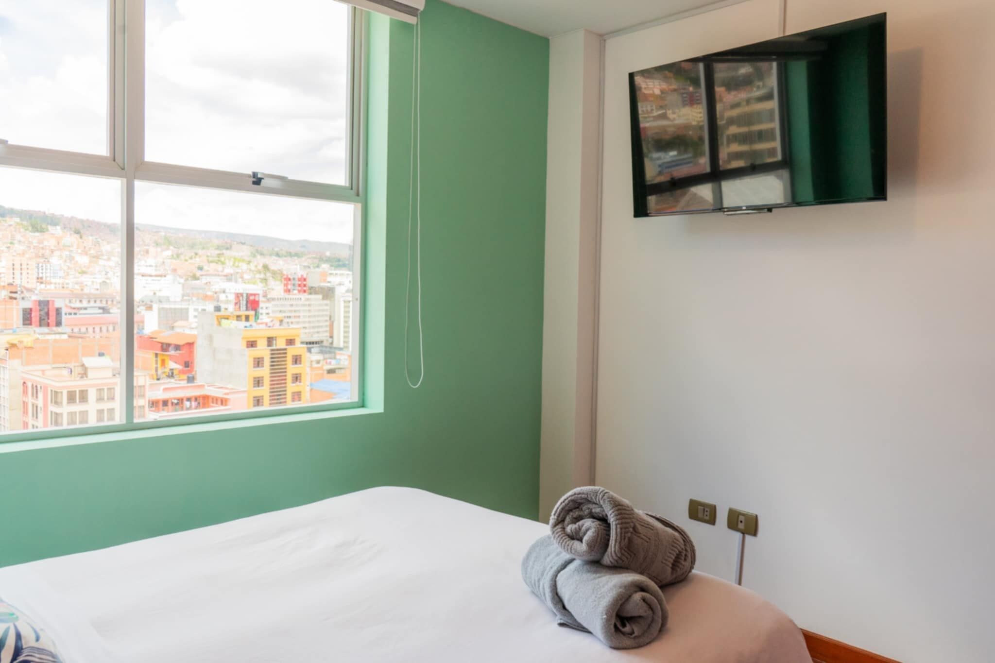 Una camera d'albergo con un letto, una finestra che si affaccia sulla città e una TV fissata al muro.