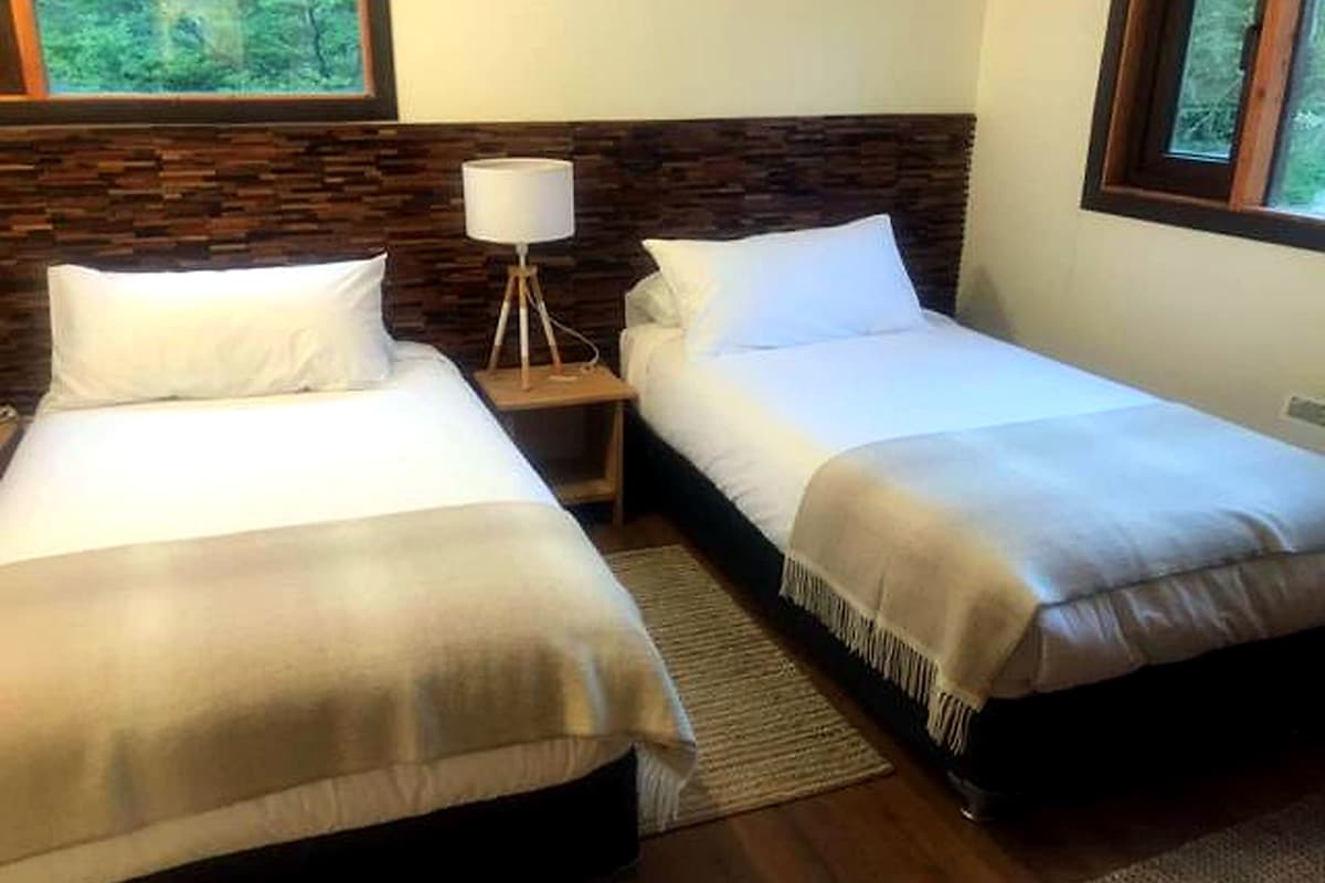 A hotel room with two single beds, a nightstand with a lamp, and a view of the outdoors through a window.