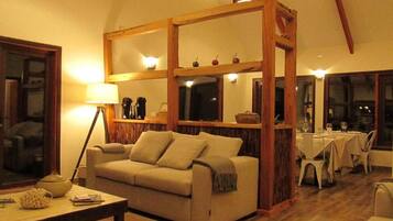 A living room with a sofa, a coffee table, a dining area with a table and chairs, and a wooden partition with shelves.