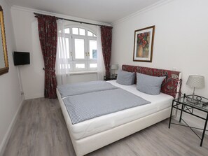 A bedroom with a large bed, a television on the wall, a window with curtains, a painting, and a bedside table with a lamp. - 7 Residence Bismarck (Sylt)