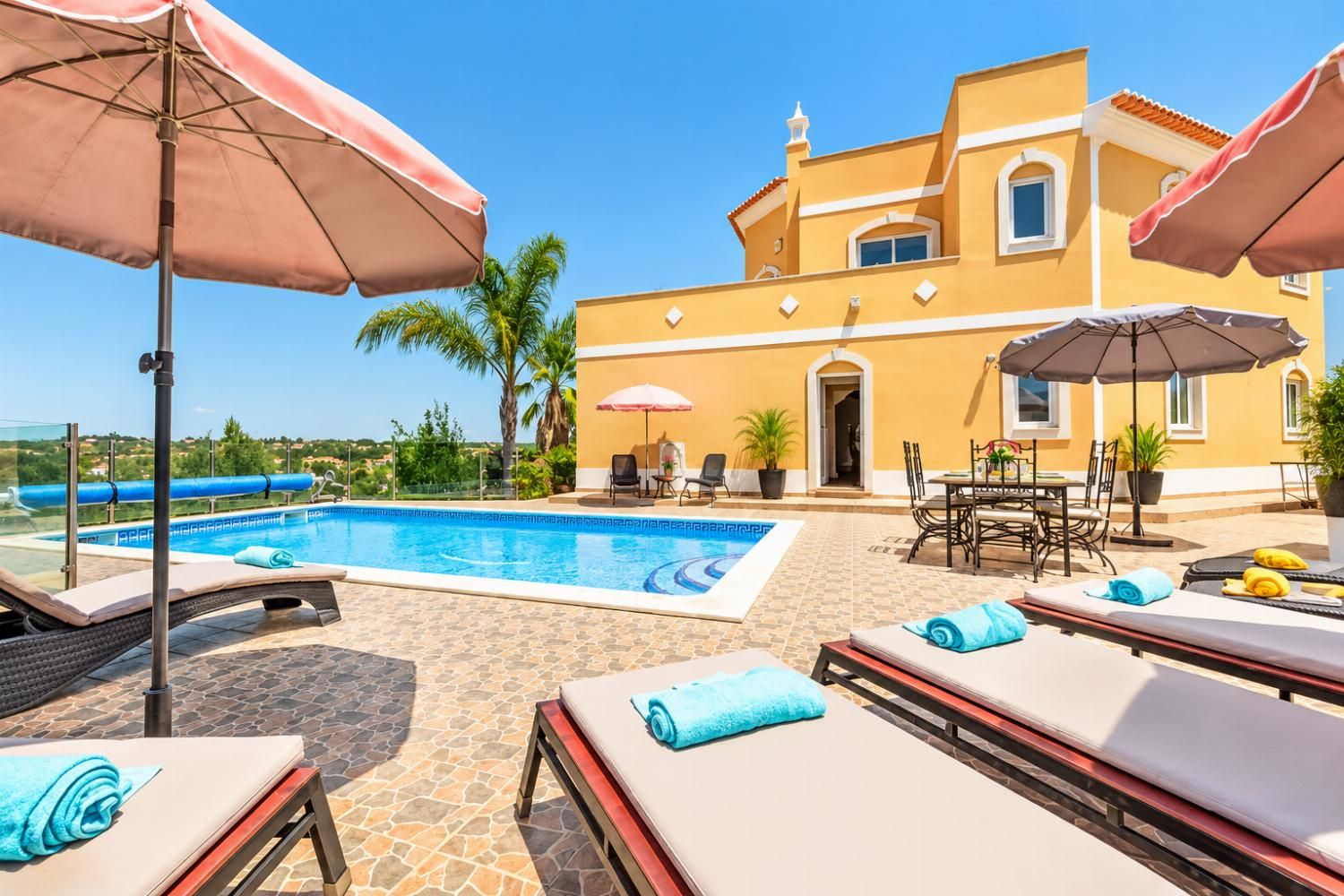 A poolside area with lounge chairs and umbrellas, a pool, and a building with a yellow facade.