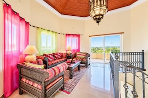 Un salon comprenant un ensemble de canapés, une table basse et un tapis à motifs. Il y a un balcon avec une balustrade et une vue sur le paysage.