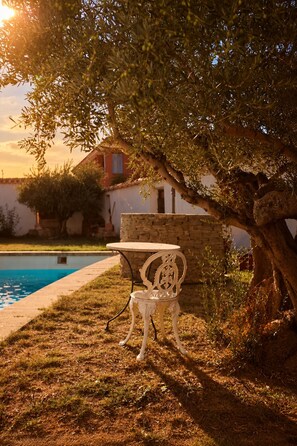 A poolside area with a white metal table and chair, a stone wall, and a tree providing shade.