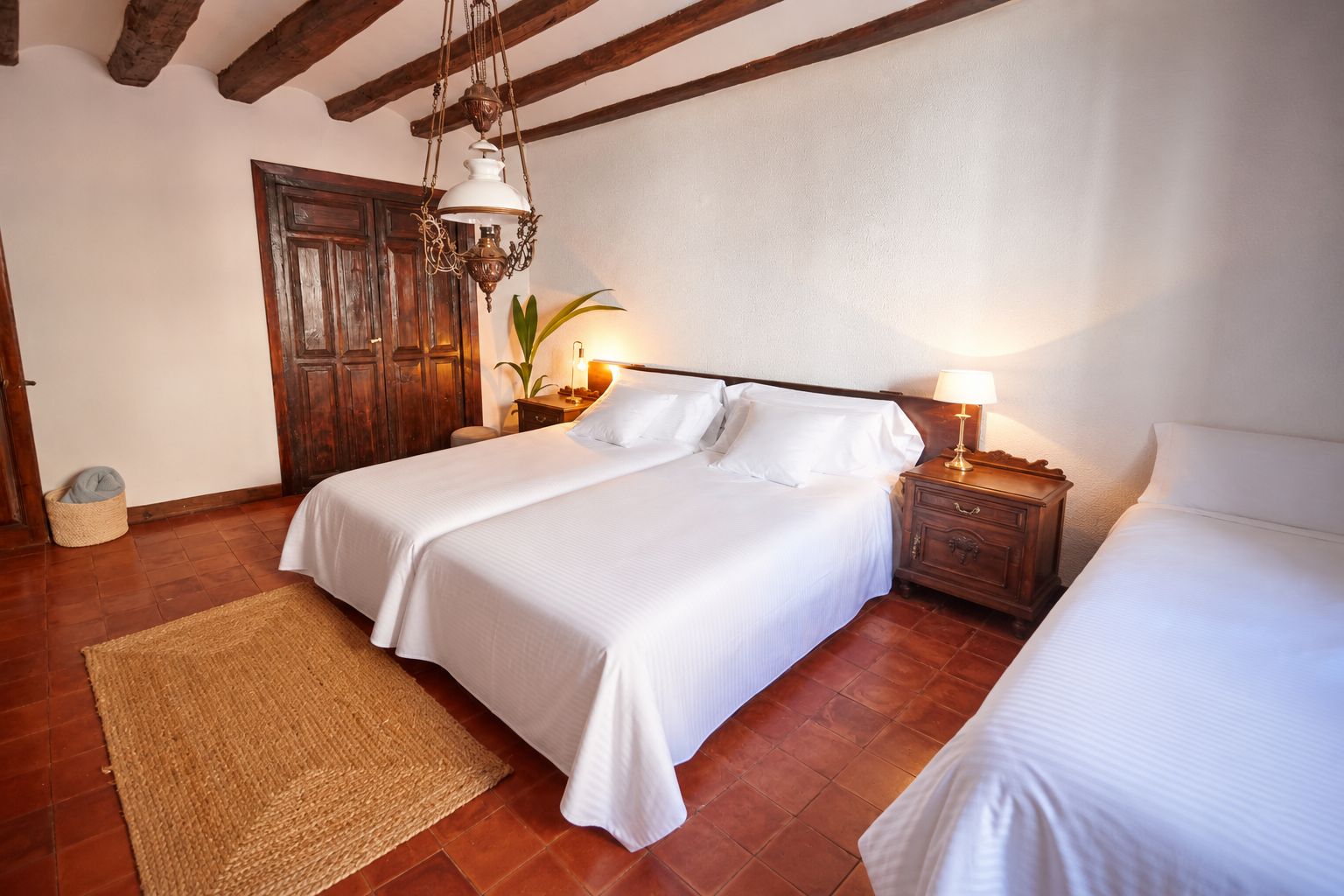 Ein Doppelbett mit weißer Bettwäsche, ein Nachttisch aus Holz mit einer Lampe und ein gewebter Teppich auf Fliesen.