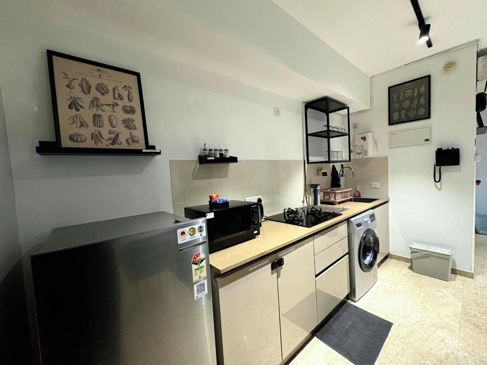 A compact kitchen with a refrigerator, microwave, stove, and washing machine. There is a framed botanical print on the wall and a shelf with kitchen items.