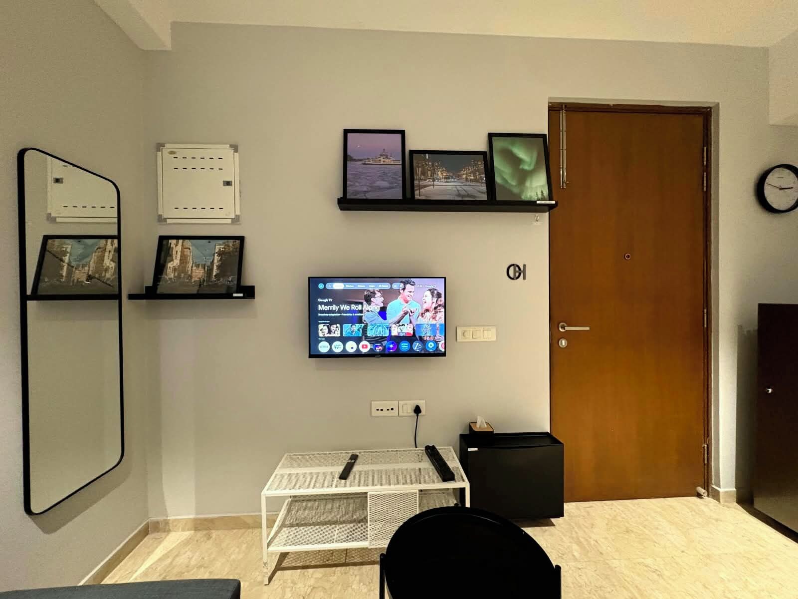 A living room with a flat-screen TV mounted on the wall, a small white table with a black chair, and a wooden door.