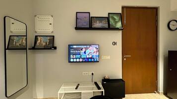 A living room with a flat-screen TV mounted on the wall, a small white table with a black chair, and a wooden door.