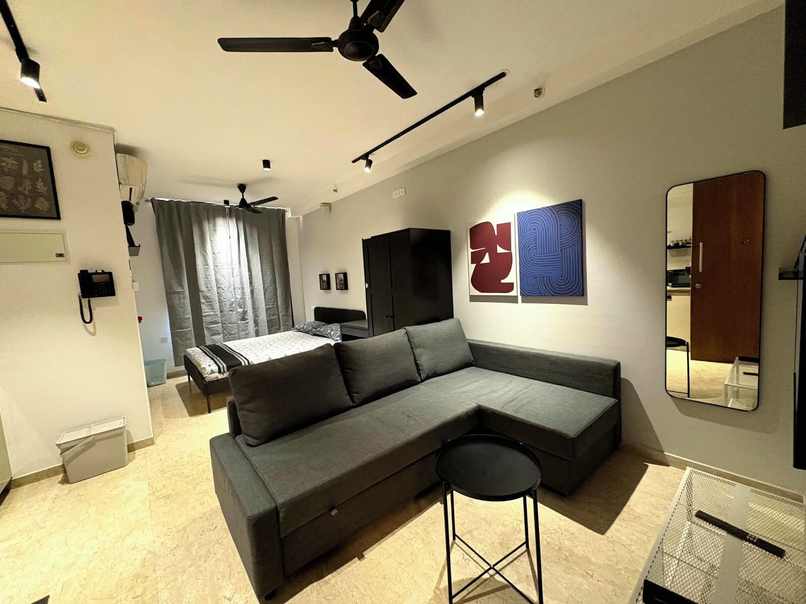 A modern living room with a sectional sofa, a small stool, a ceiling fan, and a bed in the background.