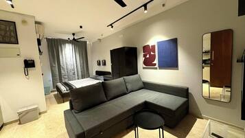 A modern living room with a sectional sofa, a small stool, a ceiling fan, and a bed in the background.