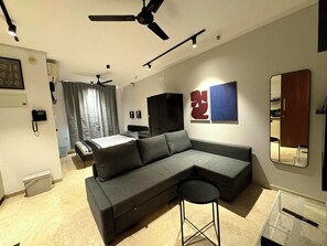Una sala moderna con un sofá seccional, un taburete pequeño, un ventilador de techo y una cama al fondo.