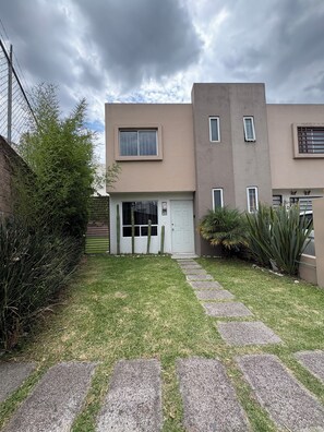 A two-story residential building with a front door, a small garden with a pathway, and a cloudy sky.