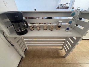 A kitchen counter with a coffee maker, a shelf with coffee mugs, and a window with a view of the sky.