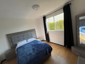 A bedroom with a bed, a nightstand, a dresser, a window with curtains, and a ceiling light.