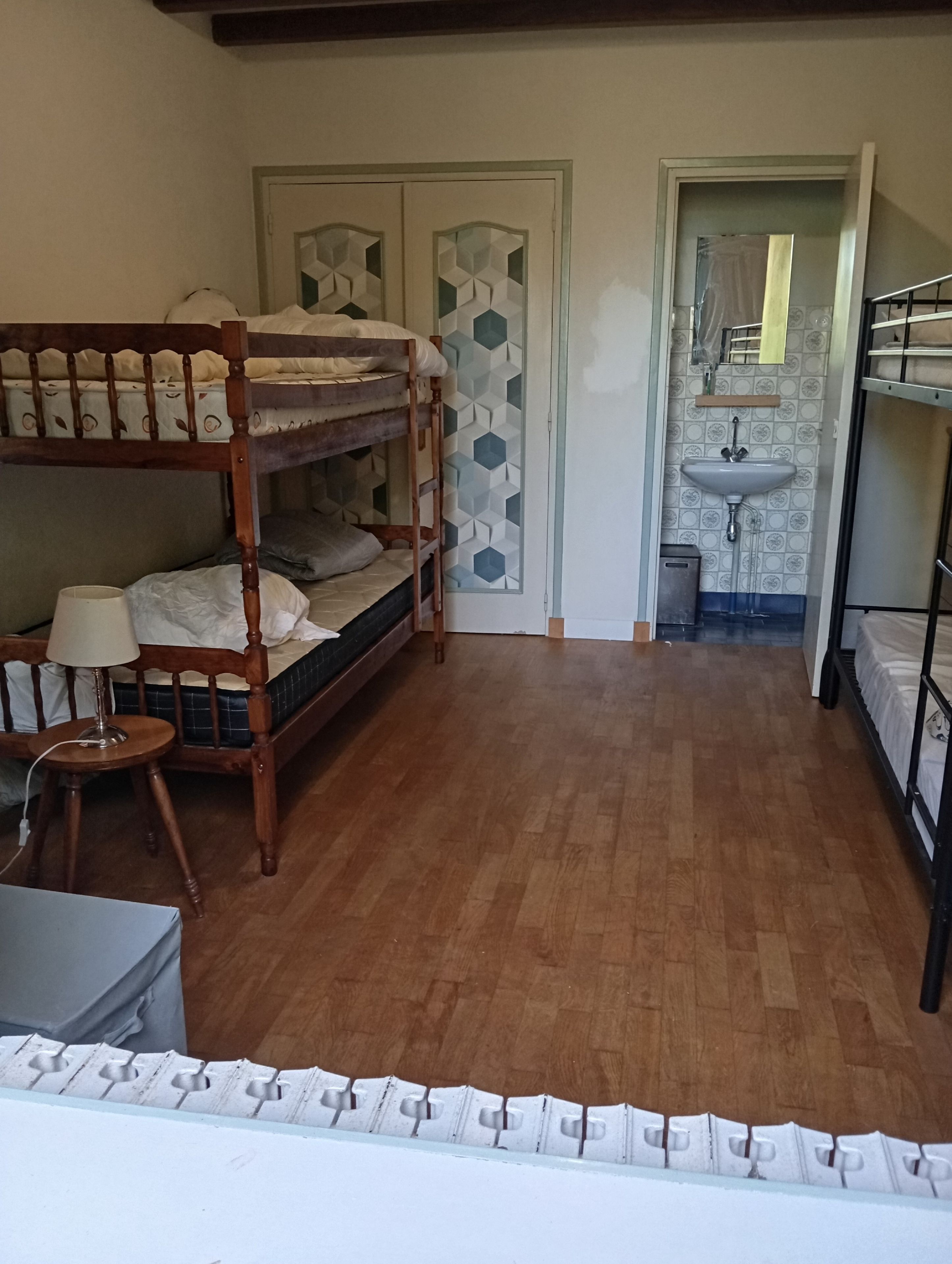 Una habitación con dos literas, piso de madera, una mesita con lámpara y una puerta que da a un baño con lavamanos.