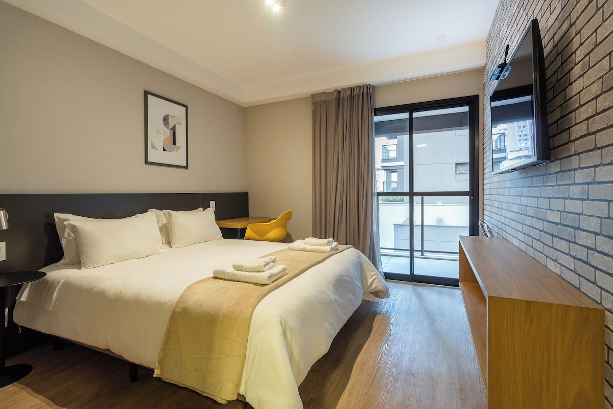 A hotel room with a large bed, a desk, a chair, a TV mounted on the wall, and a window with a view of the outside.