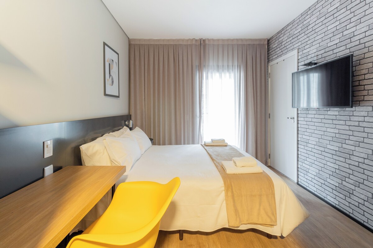 A hotel room with a bed, a wooden desk, a yellow chair, a flat-screen TV mounted on a brick wall, and a window with curtains.