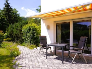 A patio with a table and chairs, a glass door leading to a garden, and a white building with a striped awning. - Holiday Apartment With 1 Bedroom (Willingen)