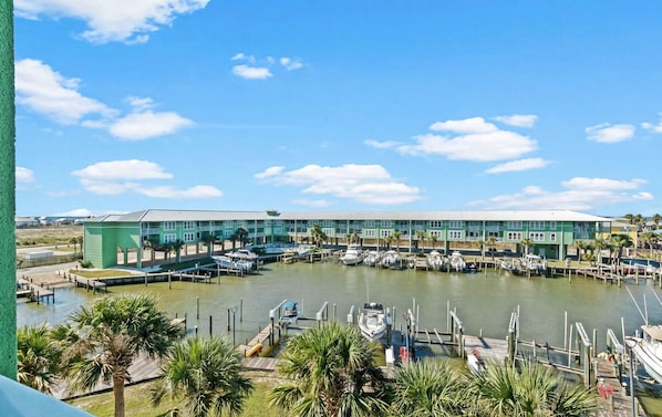 A marina with multiple docks and boats, a large building with a flat roof, and palm trees in the foreground.