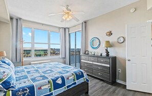 A bedroom with a bed, a dresser, a ceiling fan, a window with a view, and a door.