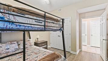 A bunk bed room with a nautical-themed bedspread, a bedside table with a lamp, and a door leading to another room.