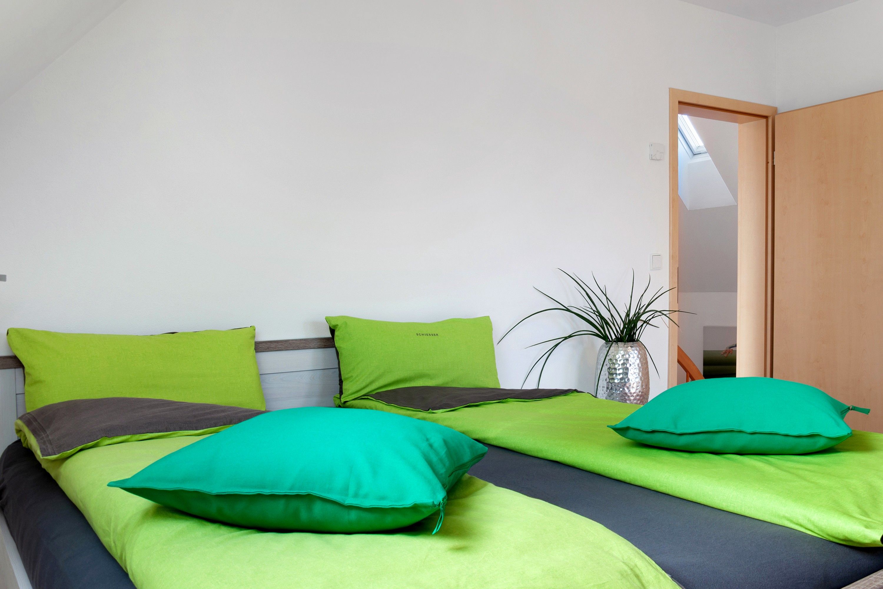 Un dormitorio con dos camas, cada una con ropa de cama verde y verde oscuro, una planta en maceta y una puerta de madera.