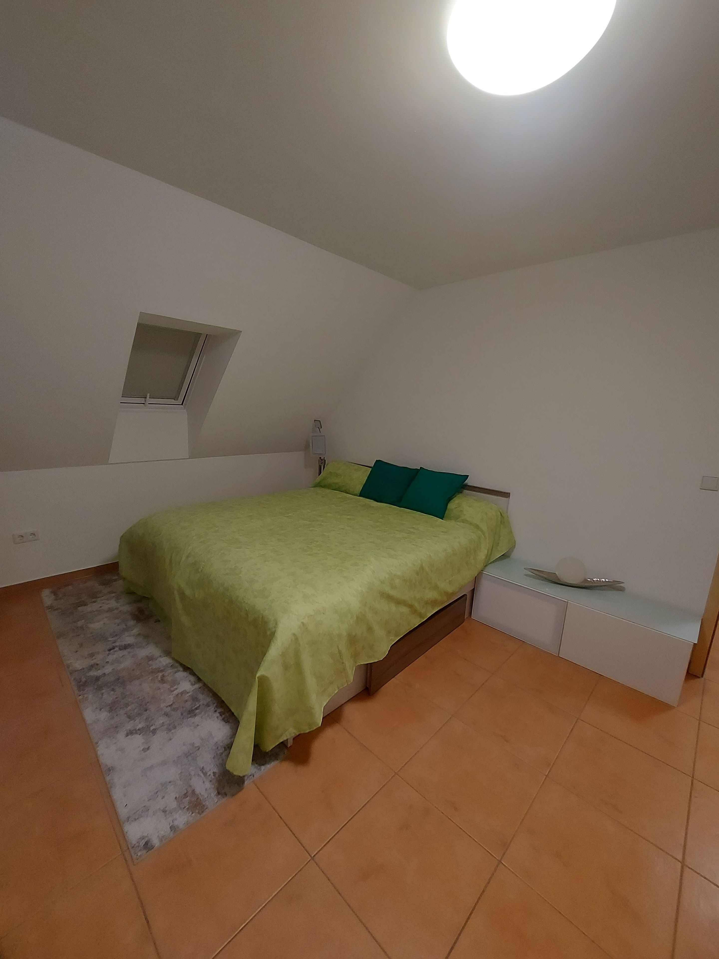 Ein Schlafzimmer mit einem Bett, einem Nachttisch, einer Lampe und einem kleinen Fenster.