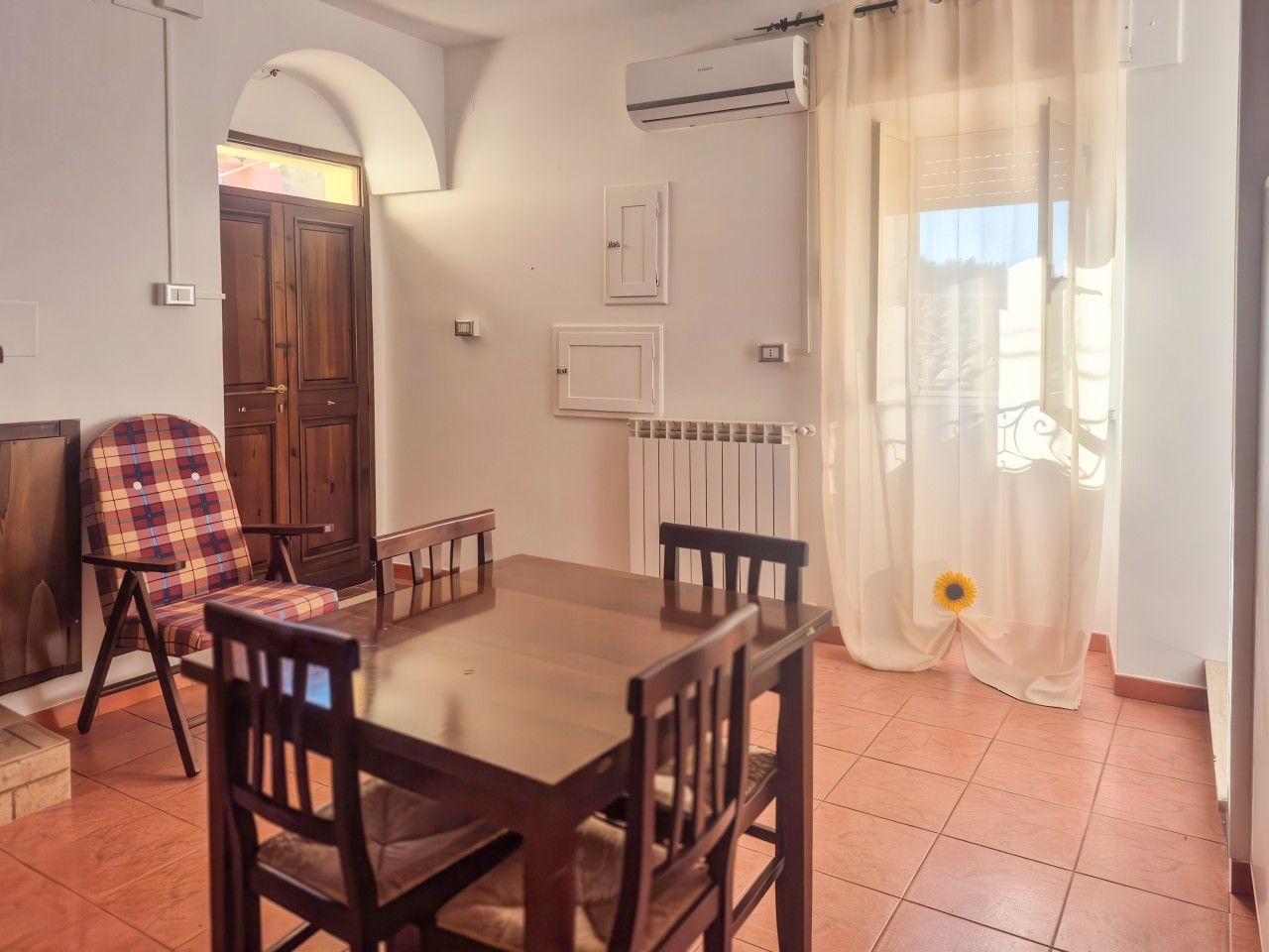 Zona pranzo con tavolo e sedie in legno, una poltrona con motivo decorativo e una finestra con tende leggere.