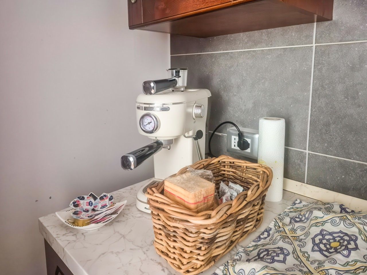 Une cuisine équipée d’une machine à café, d’un panier de pain et d’une nappe à motifs.
