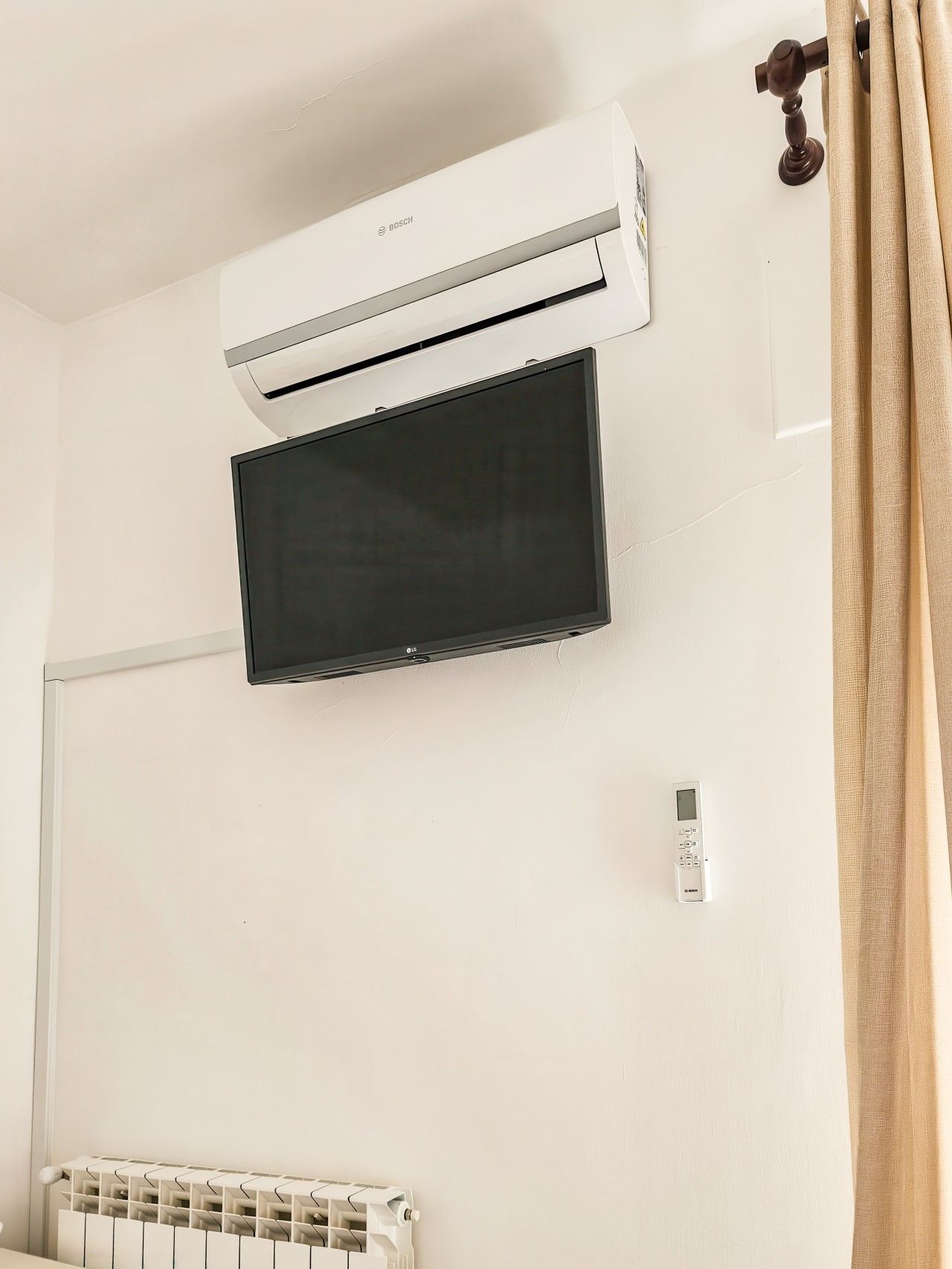 A room with a white wall, a mounted flat-screen TV, a wall-mounted air conditioner, a thermostat, and a radiator.