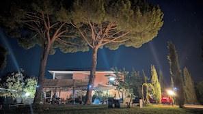 Scena notturna all'aperto con una zona salotto coperta, alberi e un edificio sullo sfondo.