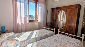 Ein Schlafzimmer mit zwei Betten, einem Holzschrank, einem Fenster mit Vorhängen und einer blauen Lampe auf einem Nachttisch.
