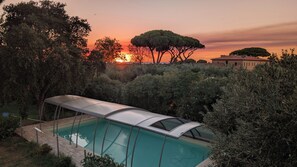 Una piscina con zona cubierta, rodeada de abundante vegetación y un atardecer de fondo.