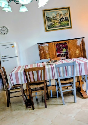 Un comedor con una mesa dispuesta para cuatro personas, sillas de madera, un refrigerador, un reloj y un cuadro de un paisaje en la pared.
