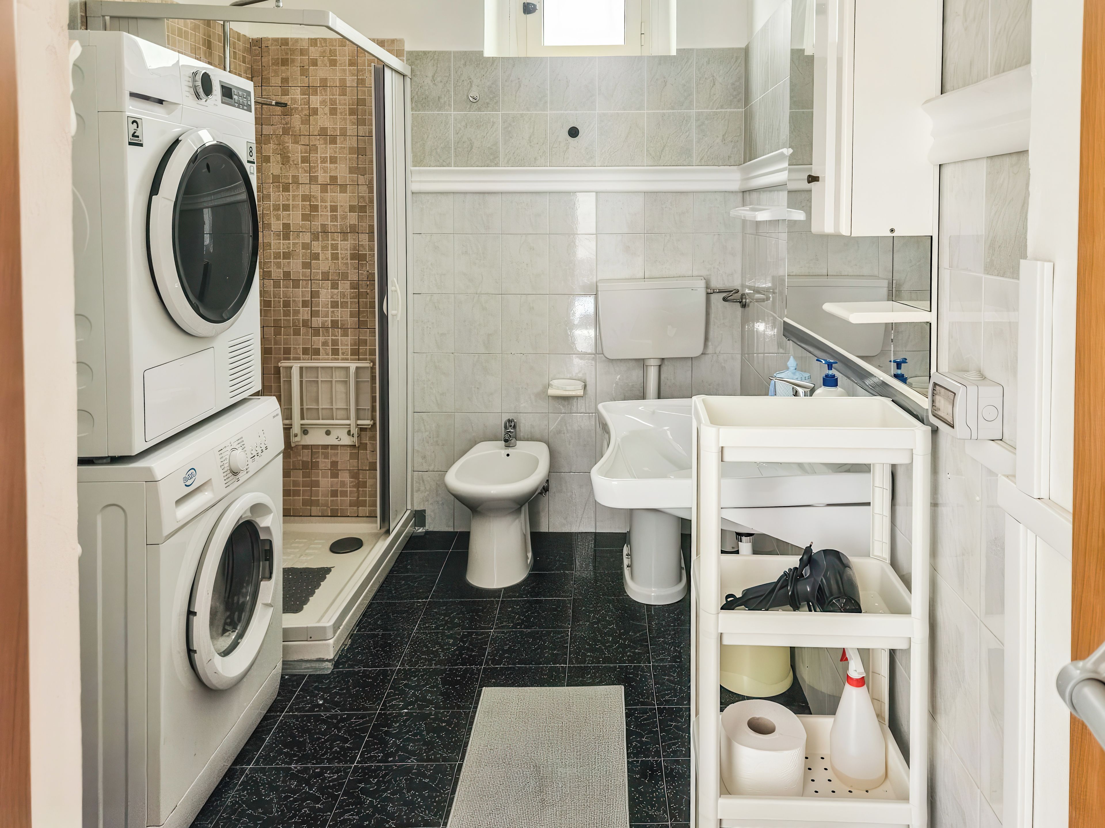 Casa de banho compacta com máquina de lavar roupa, bidé, lavatório e uma pequena estante com produtos de higiene pessoal.