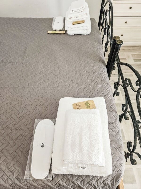 Una cama bien hecha con colcha gris, almohada blanca y toalla blanca doblada con una etiqueta.